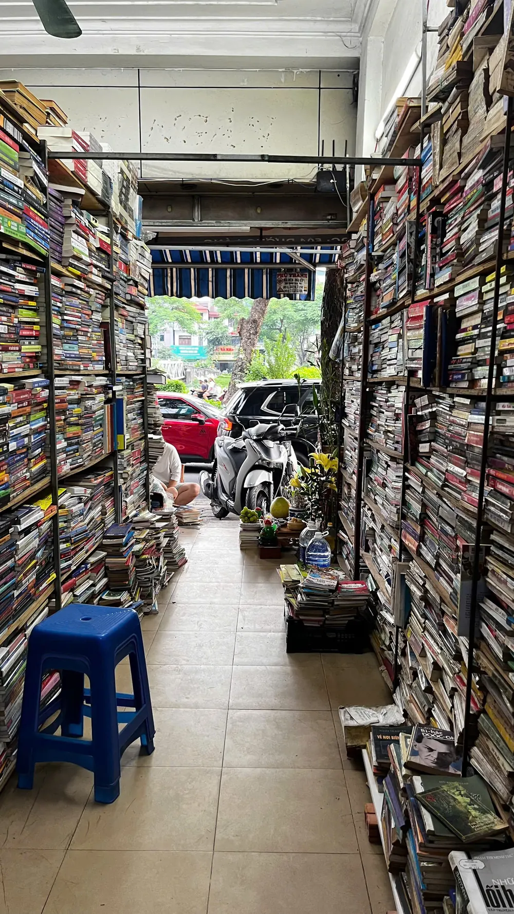 A narrow aisle in a secondhand bookshop, with used books stacked floor to ceiling on both sides and a blue plastic stool in the foreground.